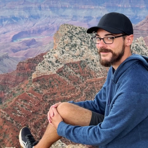 Josh Vickerson is sitting in front of the Grand Canyon, wearing a black baseball cap and navy blue sweatshirt. He has a dark, medium-length beard, glasses and is smiling.