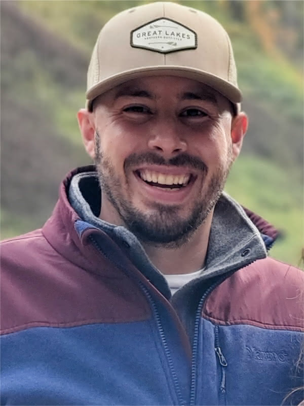 Josiah Peterham, a white man with a short dark beard, wearing a tan baseball cap, and a red and blue jacket smiling.